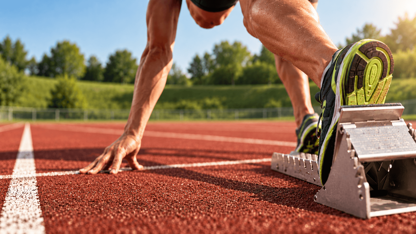 Sprintstart auf einer roten Tartanbahn: Hand auf der Bahn, Schuh am Startblock und grüner Wall im Hintergrund.